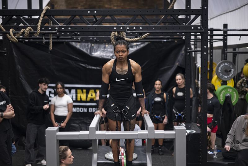 Shernelle on the dip bars wearing a weighted belt, focused before a rep, with other competitors watching.