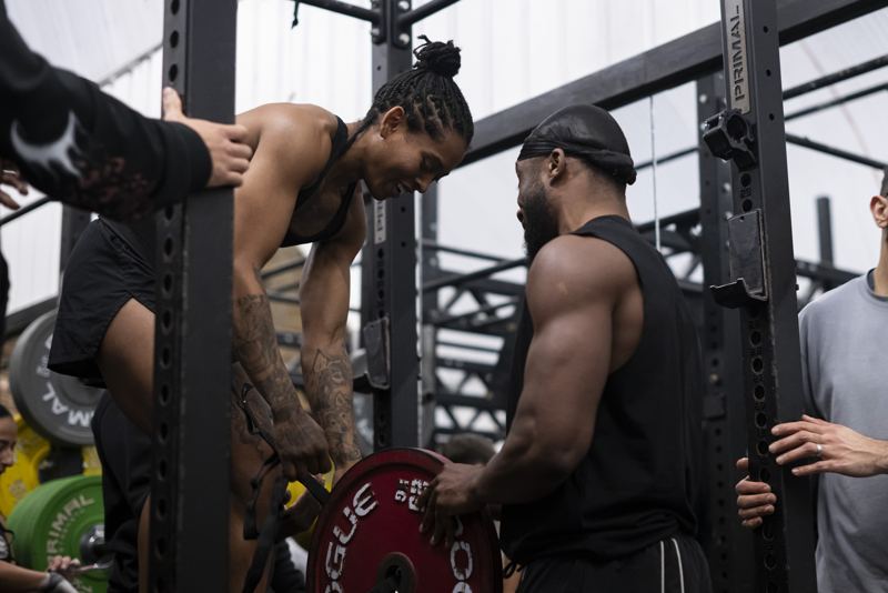 Shernelle smiling as she sets up a weight plate on her belt between attempts, with another athlete helping.